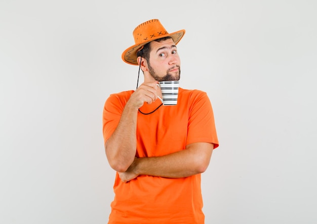 Jonge man houdt kopje drank in oranje t-shirt, hoed en ziet er verstandig uit, vooraanzicht.