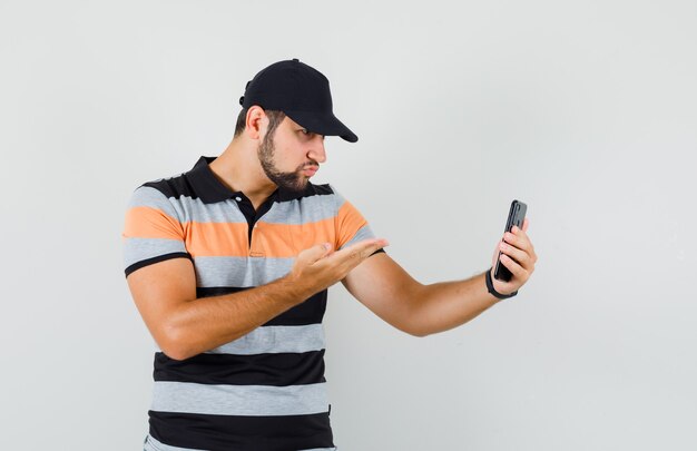 Jonge man iets bespreken op videochat in t-shirt, pet en op zoek verward. vooraanzicht.