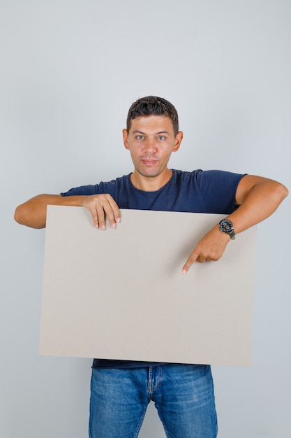 Jonge man iets op poster in blauw t-shirt, spijkerbroek en op zoek positief, vooraanzicht.