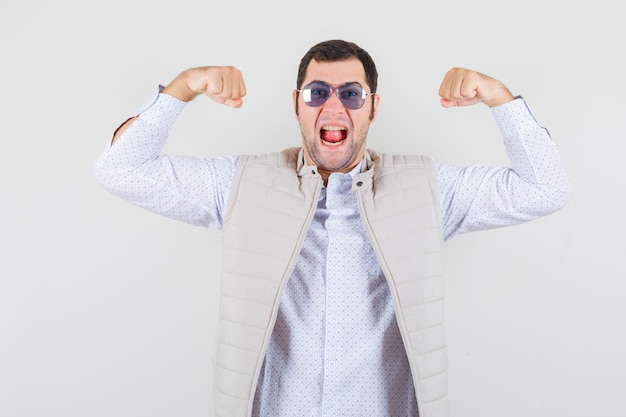 Jonge man in beige jas en pet op bril terwijl de winnaar pose toont, tong uitsteekt en op zoek optimistisch, vooraanzicht.