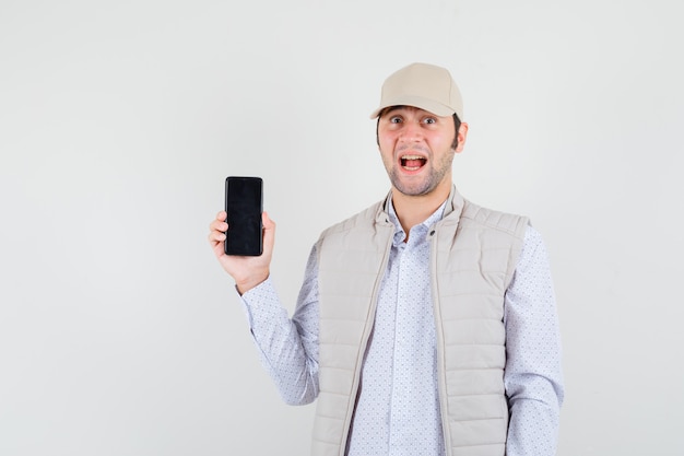 Jonge man in beige jasje en pet die telefoon tonen en verbaasd, vooraanzicht kijken.