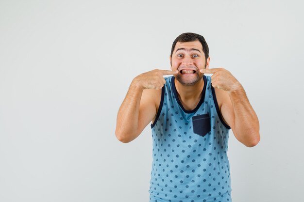 Gratis foto jonge man in blauw hemd wijzend op zijn tanden en ziet er vrolijk uit