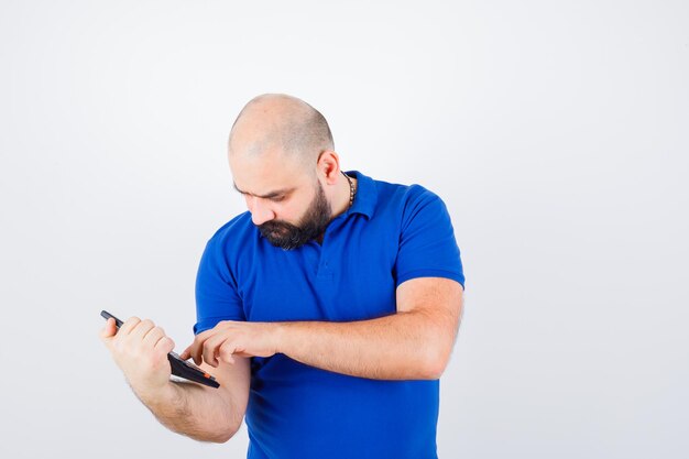 Jonge man in blauw shirt berekent via rekenmachine en kijkt gefocust, vooraanzicht.