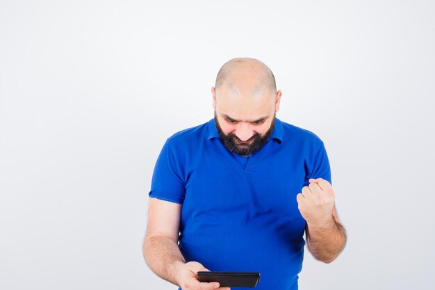 Jonge man in blauw shirt die naar de rekenmachine kijkt terwijl hij zijn vuist opheft en er vrolijk uitziet, vooraanzicht.
