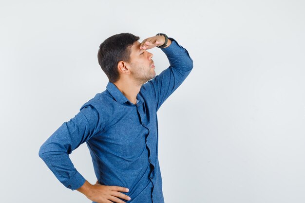 Jonge man in blauw shirt die ver weg kijkt met de hand boven het hoofd, vooraanzicht.