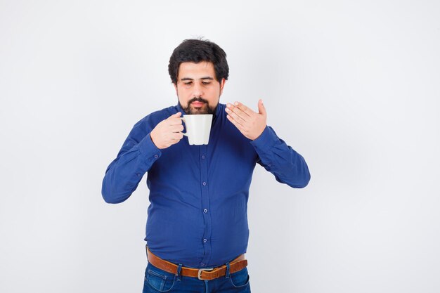 Jonge man in blauw shirt en spijkerbroek die een kopje water probeert te drinken en er serieus uitziet, vooraanzicht.