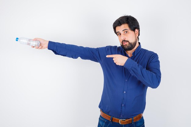 Jonge man in blauw shirt en spijkerbroek die een waterfles vasthoudt en erop wijst en er serieus uitziet, vooraanzicht.