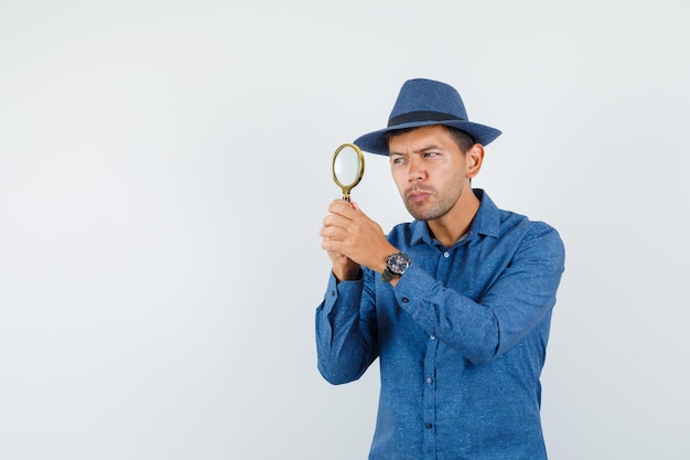 Gratis foto jonge man in blauw shirt, hoed kijkend door vergrootglas, vooraanzicht.