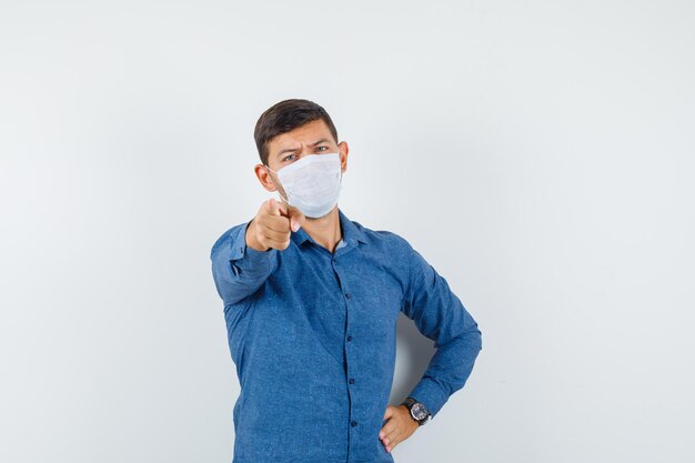 Jonge man in blauw shirt, masker wijzend op camera en serieus kijkend, vooraanzicht.