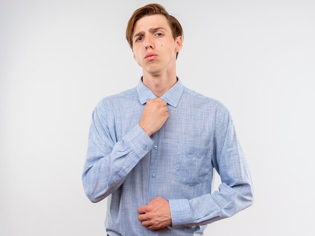 Jonge man in blauw shirt met zelfverzekerde uitdrukking die zijn kraag bevestigt die zich over witte muur bevindt