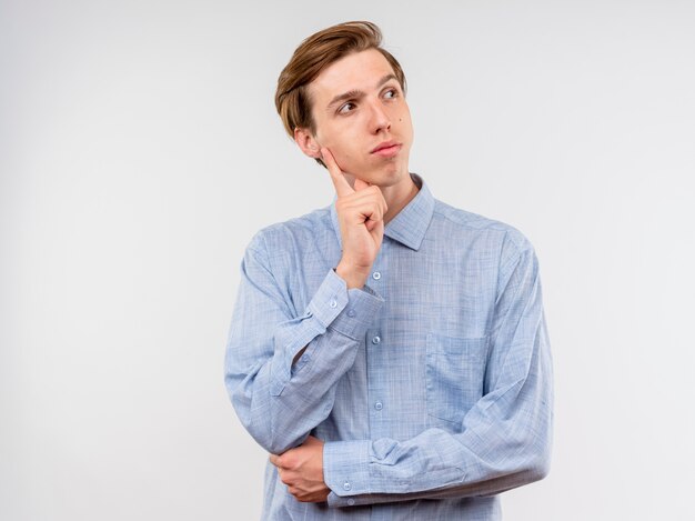 Jonge man in blauw shirt opzij kijken met peinzende uitdrukking denken staande op witte achtergrond