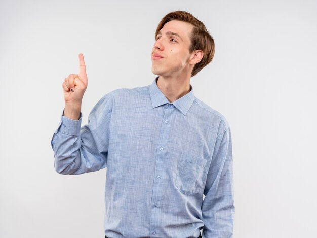 Jonge man in blauw shirt opzij wijzend naar iets met wijsvinger glimlachend zelfverzekerd staande op witte achtergrond