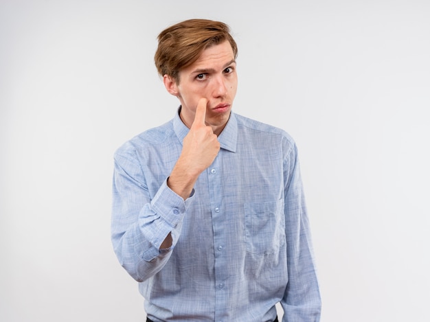 Jonge man in blauw shirt wijzend met index figner naar zijn oog staande over witte muur