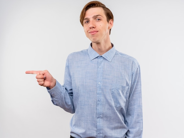 Jonge man in blauw shirt wijzend met wijsvinger naar de zijkant glimlachend zelfverzekerd staande over witte muur