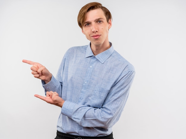 Jonge man in blauw shirt wijzend met wijsvingers naar de zijkant glimlachend zelfverzekerd staande over witte muur