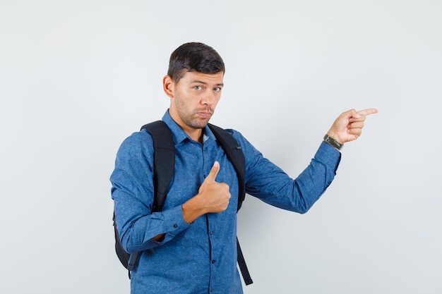 Jonge man in blauw shirt wijzend naar de zijkant met duim omhoog, vooraanzicht.