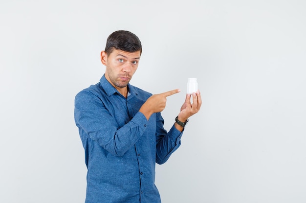 Jonge man in blauw shirt wijzend op fles pillen en aarzelend, vooraanzicht.