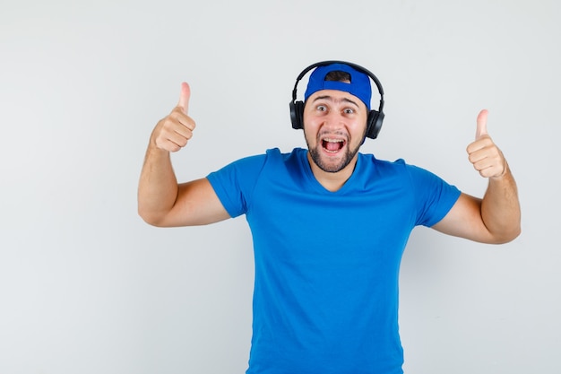 Jonge man in blauw t-shirt en pet met koptelefoon terwijl hij duimen opdagen en er gelukkig uitziet