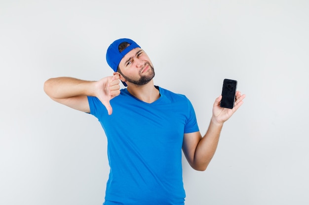 Jonge man in blauw t-shirt en pet met mobiele telefoon met duim naar beneden en ontevreden op zoek