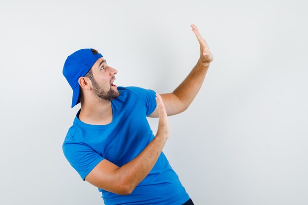 Jonge man in blauw t-shirt en pet met stopgebaar en bang op zoek
