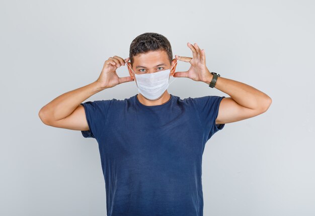 Jonge man in donkerblauw t-shirt met medische masker en op zoek voorzichtig, vooraanzicht.