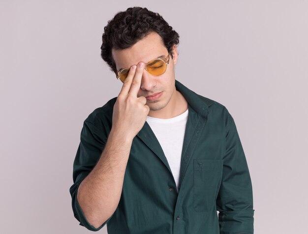 Jonge man in een groen shirt met bril aanraken van zijn voorhoofd op zoek moe en verveeld staande over witte muur