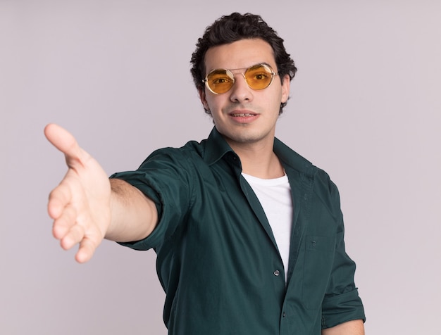 Jonge man in een groen shirt met bril op zoek naar de voorkant met handgroet glimlachend vriendelijk staande over witte muur