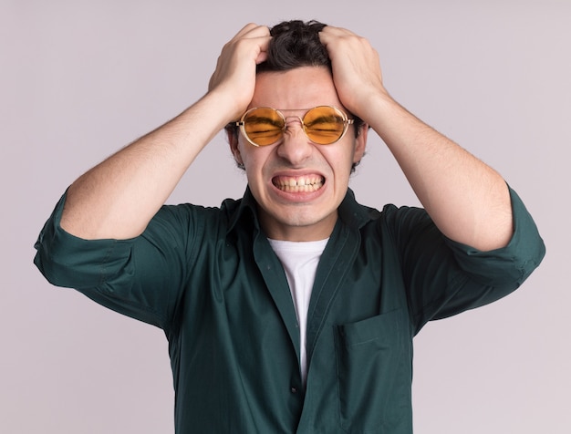 Jonge man in een groen shirt met een bril die wild gaat en zijn haar trekt dat zich over een witte muur bevindt