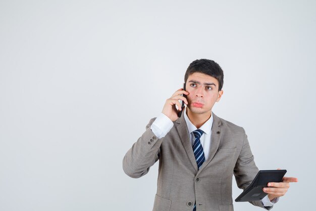 Jonge man in formeel pak die met de telefoon praat, rekenmachine vasthoudt, ergens aan denkt