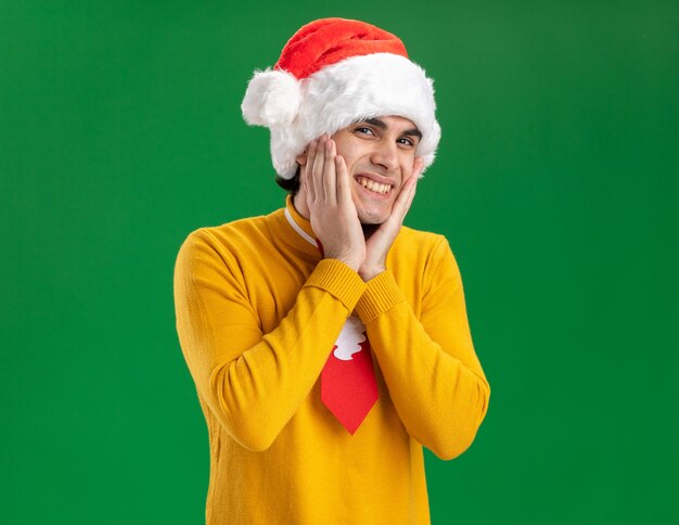 Jonge man in gele coltrui en kerstmuts met grappige stropdas gelukkig en vrolijk lachend staande over groene muur