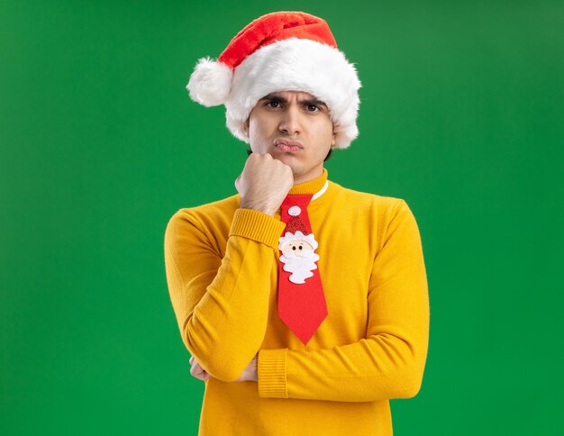Jonge man in gele coltrui en kerstmuts met grappige stropdas kijken camera ontevreden met hand op zijn kin staande over groene achtergrond