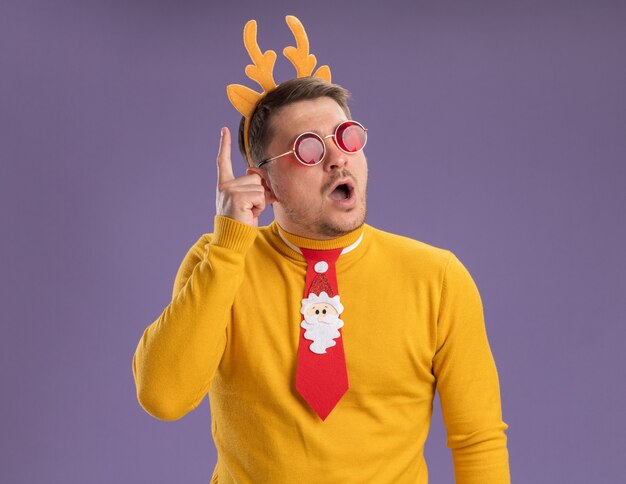 Jonge man in gele coltrui en rode bril met grappige rode stropdas en rand met herten hoorns op hoofd opzij kijken verbaasd tonen wijsvinger met nieuw geweldig idee staande over paarse achtergrond