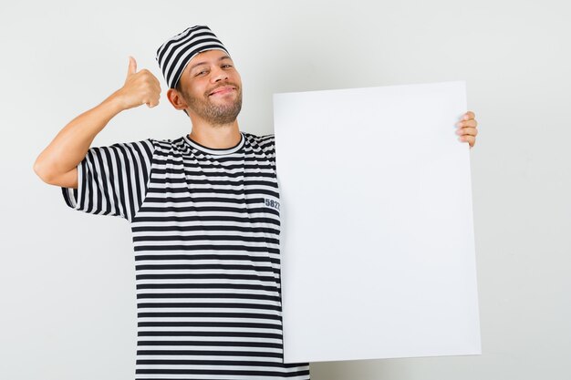Gratis foto jonge man in gestreepte t-shirthoed die duim toont die leeg canvas houdt en vrolijk kijkt