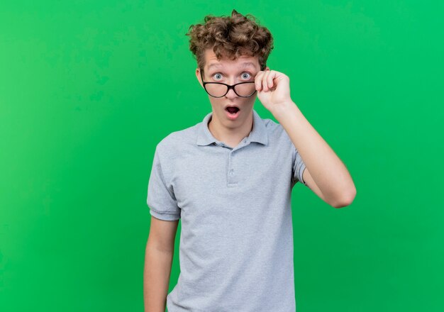 Jonge man in glazen met grijze poloshirt puuting bril af verrast en verbaasd staande over groene muur