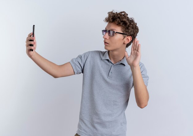 Jonge man in grijs poloshirt met smartphone doet selfie zwaaien met hand glimlachend staande over witte muur