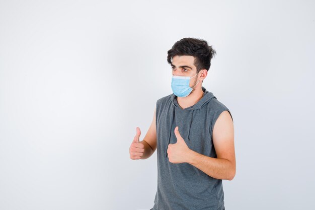 Jonge man in grijs t-shirt met masker terwijl hij zijn duimen laat zien en er serieus uitziet