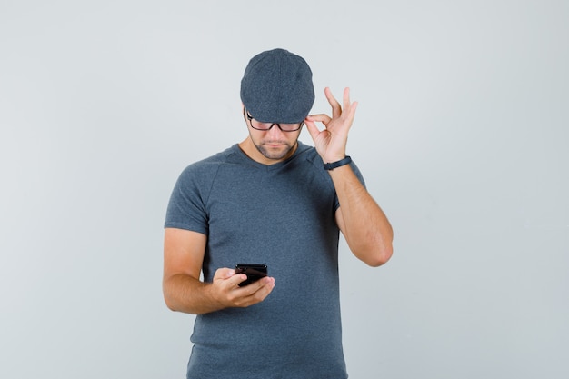 Jonge man in grijs t-shirt, pet met behulp van mobiele telefoon en op zoek naar druk, vooraanzicht.