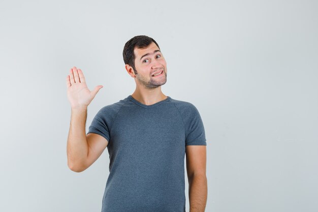 Jonge man in grijs t-shirt zwaaiende hand voor groet en op zoek vrolijk