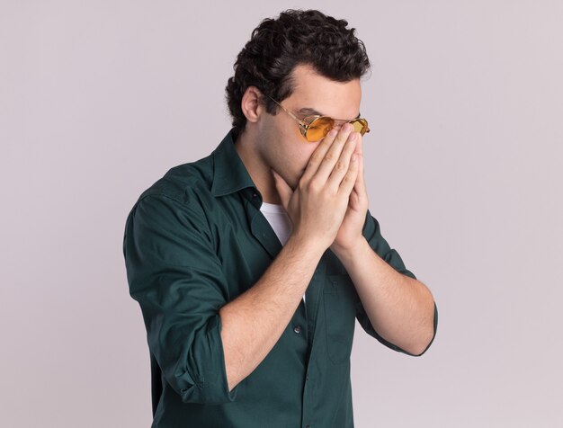 Jonge man in groen shirt met bril beklemtoond en depressief handpalmen bij elkaar te houden staande over witte muur