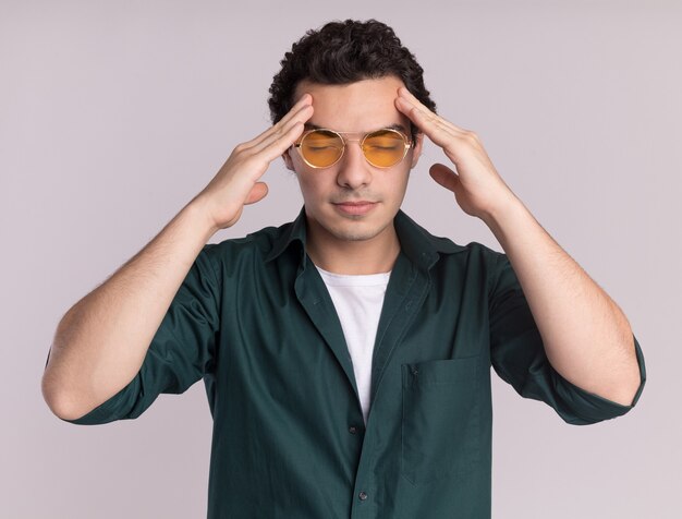 Jonge man in groen shirt met bril hoofd aanraken moe en overwerkt met hoofdpijn staande over witte muur