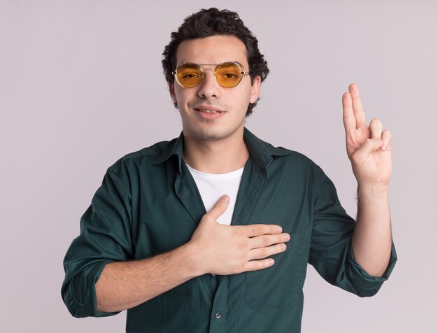 Jonge man in groen shirt met bril kijken naar voorkant vloekend met hand op borst en vingers, een loyaliteitsbelofte doen glimlachend staande over witte muur