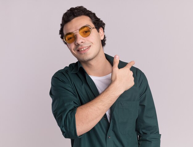 Jonge man in groen shirt met bril kijken voorkant glimlachend wijzend met vinger terug staande over witte muur