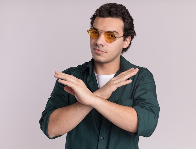 Jonge man in groen shirt met bril op zoek naar voorzijde met ernstig gezicht handen kruisen staan over witte muur