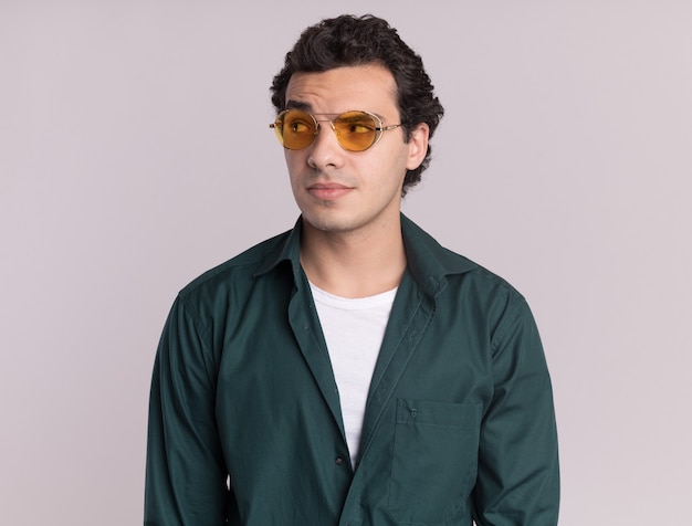 Jonge man in groen shirt met bril op zoek opzij met sceptische uitdrukking staande over witte muur