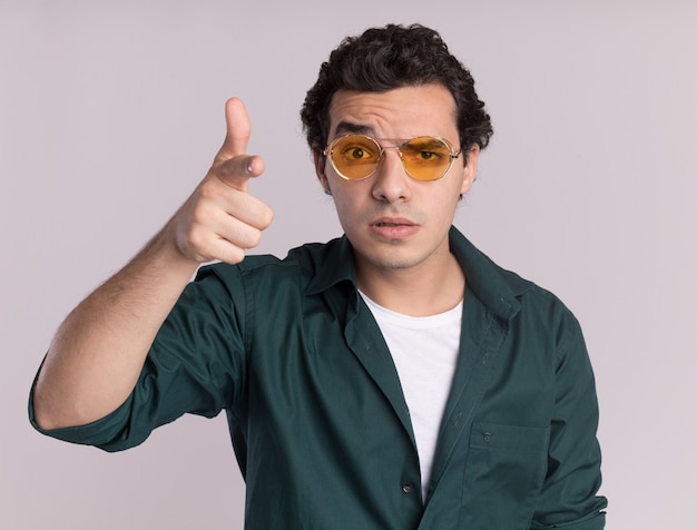 Jonge man in groen shirt met bril wijzend met wijsvinger vooraan wordt verward staande over witte muur