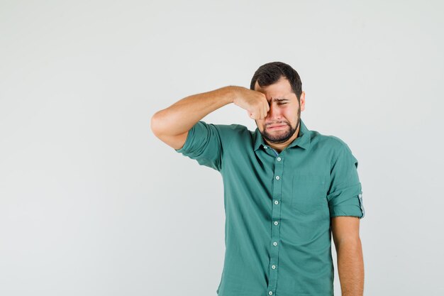 Jonge man in groen shirt wrijft in zijn oog terwijl hij huilt en boos kijkt, vooraanzicht.