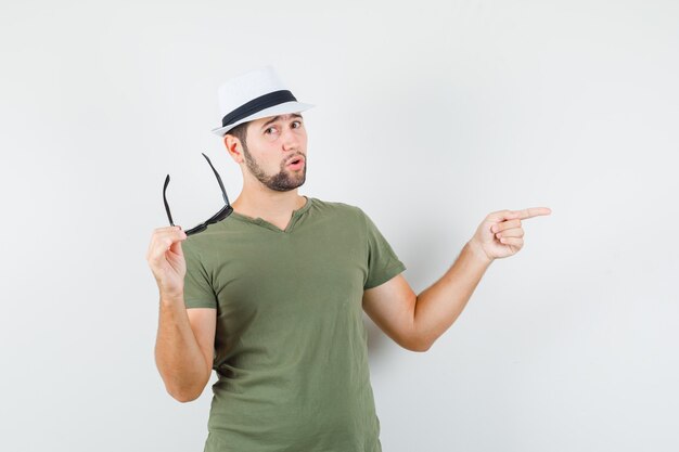 Jonge man in groen t-shirt en hoed wijst weg terwijl hij een bril vasthoudt en er cool uitziet