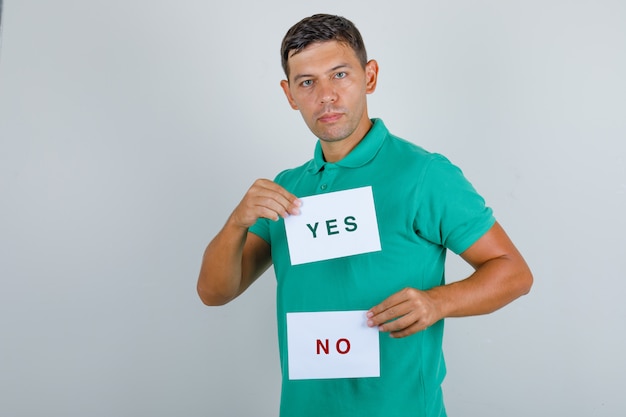 Jonge man in groen t-shirt met vellen papier met antwoorden en op zoek naar ernstige, vooraanzicht.