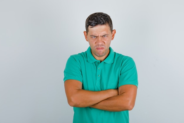 Jonge man in groen t-shirt staan met gekruiste armen en kijkt boos, vooraanzicht.