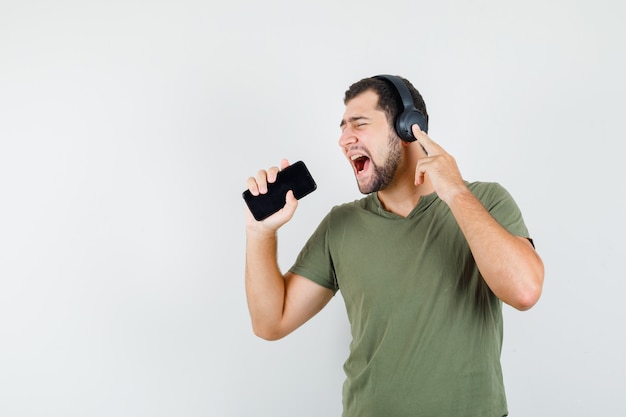 Jonge man in groen t-shirt zingen in mobiel als microfoon en op zoek komisch